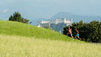 Man und Frau laufen aufwärts auf einen Wiesenweg, im Hintergrund Festung der Stadt Salzburg | © Tourismusverband Bergheim/Bryan Reinhart