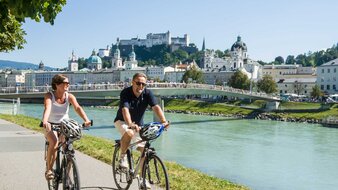 2 Radfahrer fahren am Ufer der Salzach, im Hintergrund die Festung Hohensalzburg | © Tourismus Salzburg/Bryan Reinhart