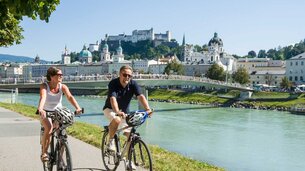 2 Radfahrer fahren am Ufer der Salzach, im Hintergrund die Festung Hohensalzburg | © Tourismus Salzburg/Bryan Reinhart