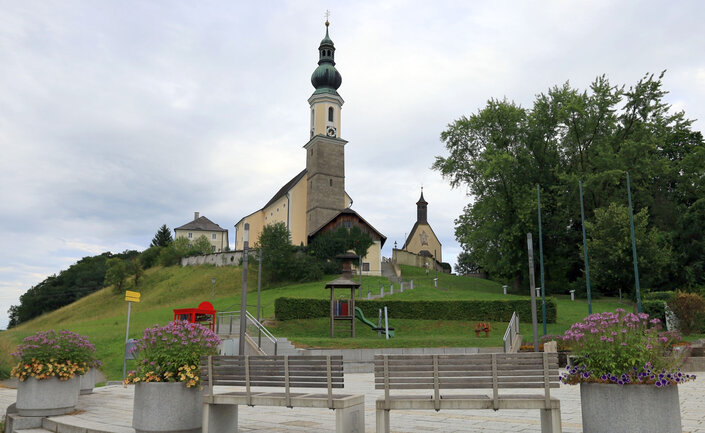 Kleiner gepflasterter Platz mit Blumentrögen und Bänken, etwas oberhalb Kirche mit Kapelle | © Tourismusverband Bergheim/Johann Taferner