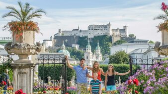 Familie (Bub und Mädchen mit Eltern) stehen bei einem eisernen Tor zu einem üppig blühenden barocken Garten, im Hintergrund die Türme des Doms und Festung  | © Tourismus Salzburg/Bryan Reinhart