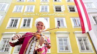 Ein als Mozart verkleideter Mann mit Geige steht vor einem gelben Gebäude | © Tourismus Salzburg