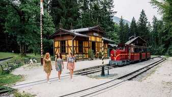 Historische rote Eisenbahn steht im Bahnhof, 3 Frauen gehen über Zuggleise | © SalzburgerLand Tourismus/Christoph Perkles