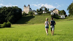 Pärchen auf Wiese und läuft Richtung Hügel mit Kapellen und Kirche | © Tourismusverband Bergheim/Bryan Reinhart